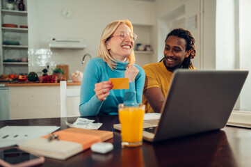 A happy multiracial couple in love is sitting at home and shopping online, using a laptop and credit card.