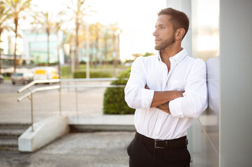 Serious businessman posing with crossed arms, looking aside at free space, standing near office building outdoors