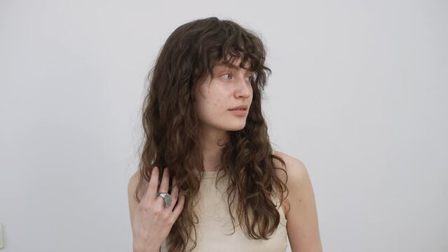 Studio portrait of attractive young woman fixing hair with hand before applying professional makeup and making stylish hairstyle, standing on isolated white background, looking at camera, slow motion.