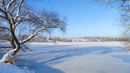 Fototapeta na wymiar In winter, in frosty and clear weather, smooth ice forms on the lake. Snow lies on the shores and trees. On the opposite shore stands a village and a mixed forest, covered in frost. Blue sky