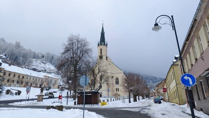 After the snowfall, snow lies on the city streets and on the roofs of the church and other buildings. The roadway has been cleared by utilities for normal traffic flow. There is frost on the trees