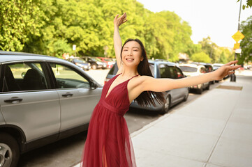 Street style outdoor portrait of Beautiful young Chinese Asian happy smiling woman walking outside sunny summer day. Attractive woman enjoys posing in front of parked cars. Big city life concept