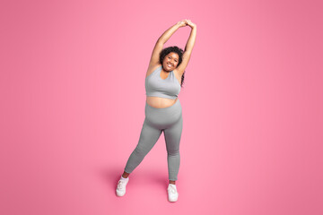 Positive black plus size lady in sportswear doing arm stretching exercises, warming up before training, pink background - obrazy, fototapety, plakaty