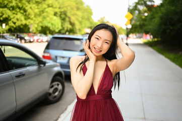 Street style outdoor portrait of Beautiful young Chinese Asian happy smiling woman walking outside sunny summer day. Attractive woman enjoys posing in front of parked cars. Big city life concept