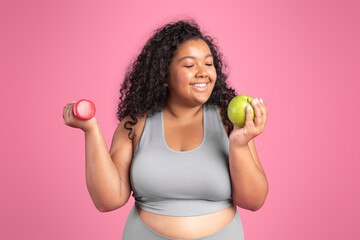 Healthy diet concept. Happy black body positive woman holding dumbbell and apple, enjoying fresh fruit and workout