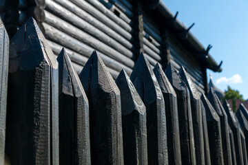 Close-up view of black wooden wall made of sharp wooden stakes standing by black log cabin in a sunny summer day. Clear blue sky. Soft focus. Copy space for your text. Protection theme.