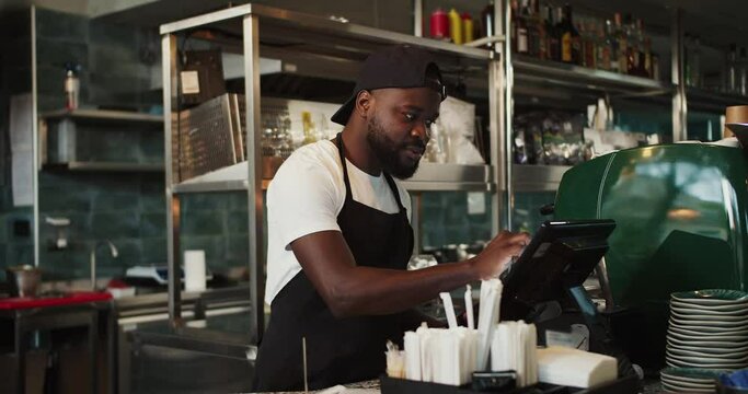 Stylish Black person in a cap - an employee of the doner market takes an order and works on an electronic guy in a cafe