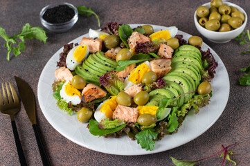 Salad with baked salmon, avocado, eggs and olives, sprinkled with sesame seeds on white plate on brown background