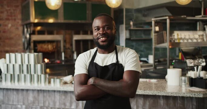A Black person in a black apron and a white T-shirt poses with arms folded on his chest against the background of the doner market