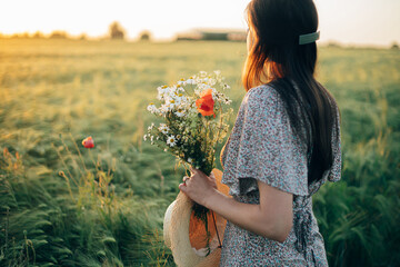 Beautiful woman with wildflowers bouquet standing in barley field in sunset light. Stylish female relaxing in evening summer countryside and gathering flowers. Atmospheric tranquil moment
