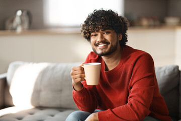 Portrait of smiling indian guy resting at home with cup of coffee