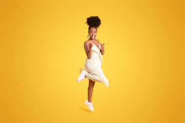 Fototapeta na wymiar Excited cheerful young african american lady in dress jumping, has fun, point fingers to camera