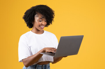 Obraz na płótnie Canvas Smiling pretty millennial black lady in casual typing on laptop, isolated on orange studio background