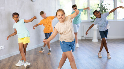 Fototapeta premium Female child performs choreographic exercises and teaches energetic mobile social dance waacking together with friends. Young girls and guys repeat movements, train in spacious studio unfocused