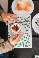 Top view of a girl's hands holding a mocha coffee with ham and cheese sandwiches on the side.