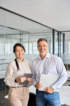 Happy Confident Professional Mature Latin Business Man And Asian Business Woman Corporate Leaders Managers Standing In Office, Two Diverse Colleagues Executives Team Together, Vertical Portrait.