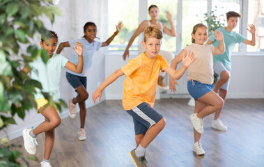 Group of positive juvenile boys and girls engaged in sport dances in training room together with young trainer