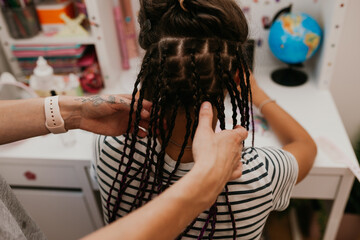 A mother braids her teenage daughter in many thin African braids while she draws on a tablet in her room.