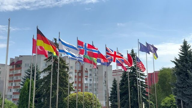 Flags of different countries of the world flutter in the wind on flagpoles against the background of green trees blue sky and houses