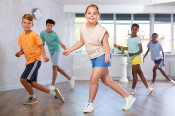 Female child performs choreographic exercises and teaches energetic mobile social dance waacking together with friends. Young girls and guys repeat movements, train in spacious studio unfocused