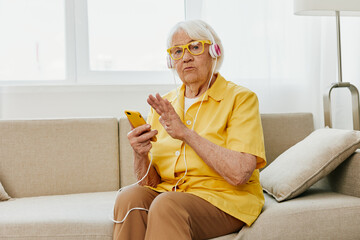 Happy elderly woman looking into her phone video call with headphones smile, technology for communication, bright modern interior, lifestyle online communication