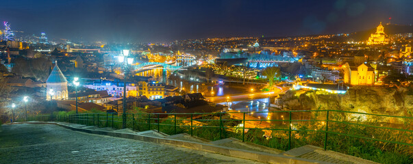 Scenic night view of Tbilisi Old town in colorful lights on banks of Kura river overlooking modern pedestrian Bridge of Peace, Georgia © JackF