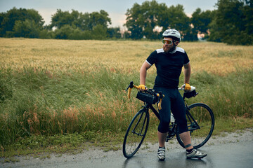 Fototapeta premium Young bearded man, cyclist in uniform, helmet and glasses sitting on bicycle on road around field background. Concept of sport, hobby, leisure activity, training, health, speed, endurance, ad