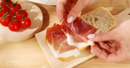 Female hands puts slices of prosciutto on cut ciabatta. Making a sandwich. Italian food. Rustic scene. Sunny day. Close up shot.