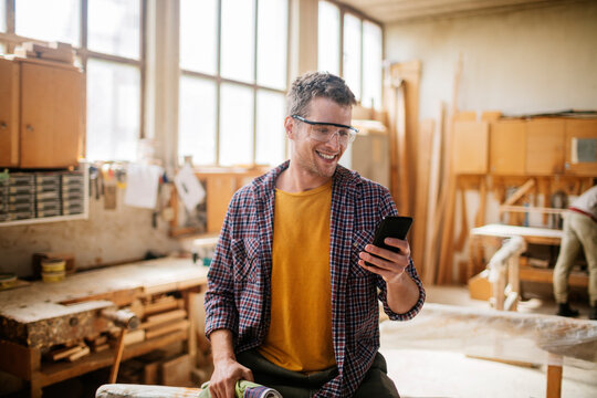 Young carpenter using a smart phone while working in his woodworking workshop