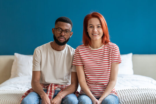 Portrait of happy just married interracial diverse couple smiling at camera while sitting together on bed at home, moved to new modern studio apartment. Harmonic family relationships concept