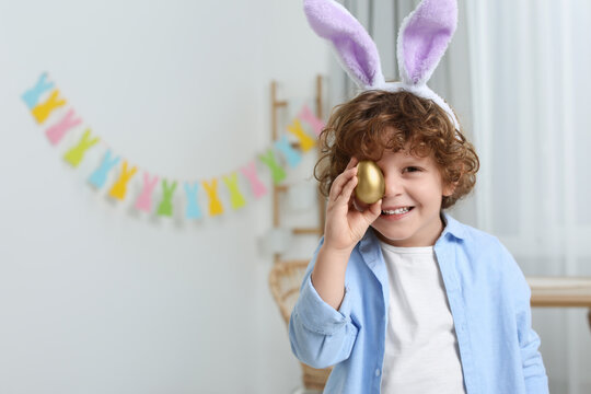 Happy boy in cute bunny ears headband covering eye with Easter egg indoors. Space for text