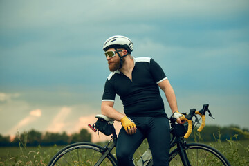 Bearded man in sportswear, glasses and helmet, sitting on bike by the road on field in chill summer evening. Concept of sport, hobby, leisure activity, training, health, speed, endurance, ad
