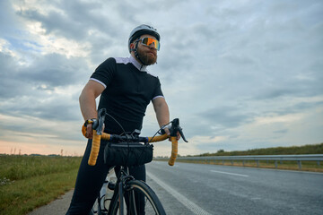 Serious, sportive bearded man in sportswear, glasses and helmet standing with bicycle by the road...