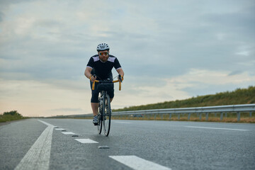 Naklejka premium Dynamic image of young, sportive, bearded man, cyclist in helmet riding bicycle on empty road in the evening. Concept of sport, hobby, leisure activity, training, health, speed, endurance, ad
