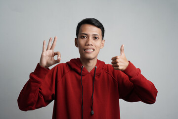 portrait of smiling young asian man wearing red hoodie showing okay sign, isolated on blue background.