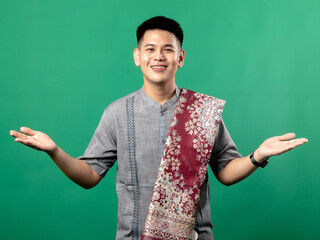 A portrait of an Indonesian Asian man wearing a gray "baju koko" (Islamic clothing) with a prayer mat on his shoulder, looking happy and posing with arms wide open, isolated on a green background.
