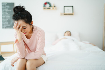 Unhappy - stressful Asian woman sitting and crying on the bed while her husband sleeping, a family and post marriage problem. Arguing Asian young couple with bad - negative relationship.