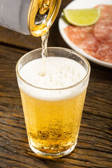 Pouring beer from a can into a glass on the table, plate of salami in the background