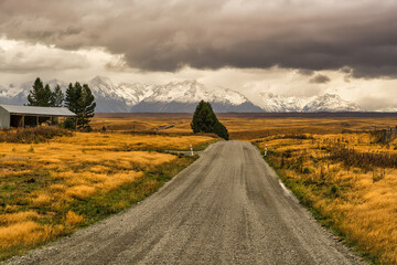 Rural country gravel road winding its way way through golden fields on a cloudy day towards the Southern Alps with a dusting of snow