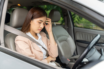 Obraz na płótnie Canvas Closed eyes tired young Asian woman with headache sitting in her parked car behind the wheel with seatbelt fasten while holding her hand over her forehead to ease the pain