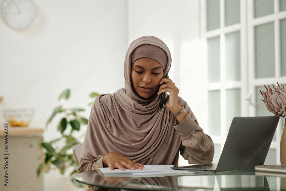 Poster young muslim woman in beige headscarf looking at document during call on smartphone while discussing