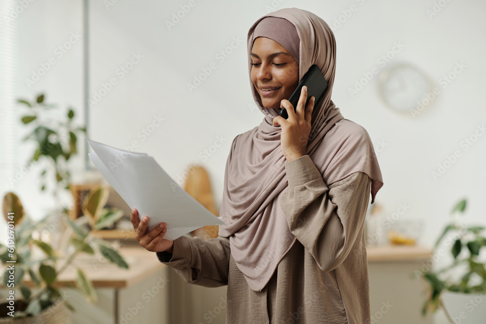 Poster young smiling businesswoman in hijab speaking on mobile phone and reading contract terms and conditi