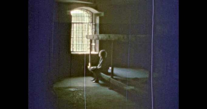Woman siting on bunk in prison. Prisoner person in confinement background of window lattice in cell, penal colony. Criminal in jail cage. Vintage color film archive. Retro, overscan archival video. 