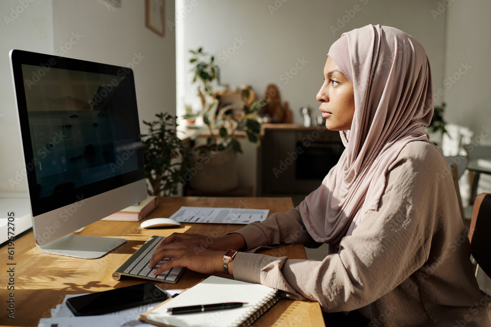 Poster Young serious Muslim woman in hijab typing on computer keyboard and looking at screen while creating new website in home environment