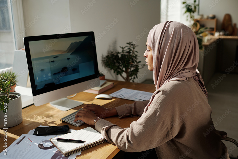 Aufkleber Young creative female webdesigner sitting by workplace in front of computer monitor and typing while working over new website