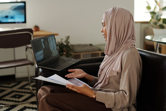 Young confident Muslim woman with financial papers looking at colleague on laptop screen while communicating in video chat at digital meeting
