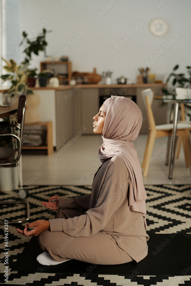 Poster side view of young muslim woman sitting in pose of lotus on mat in front of camera against home envi