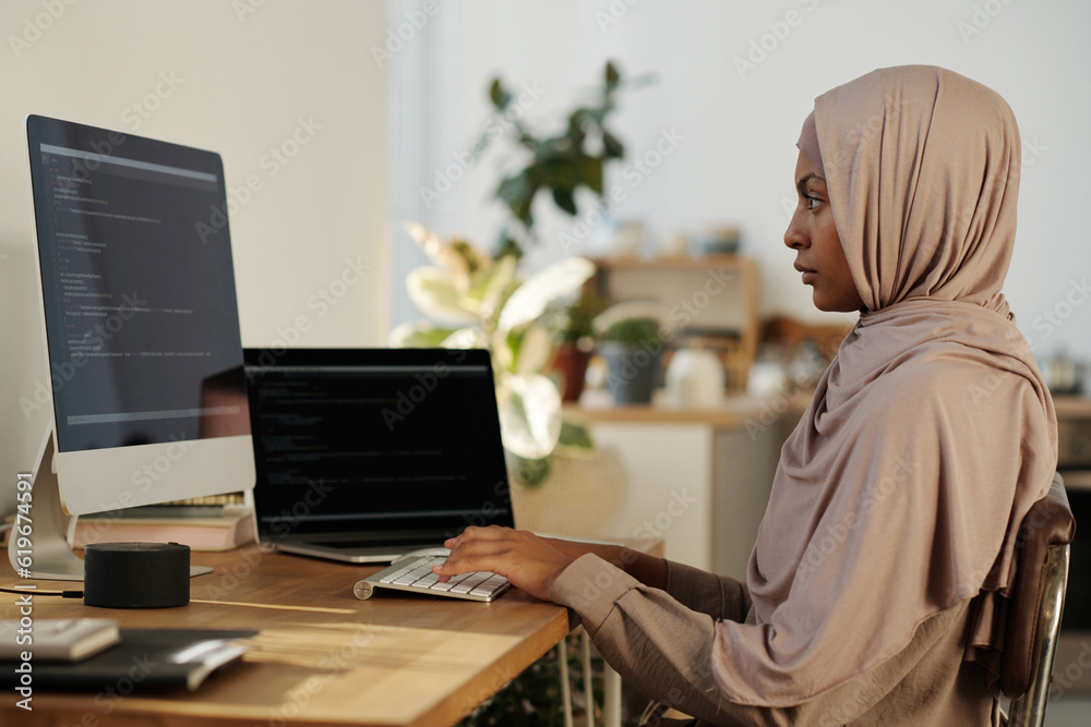 Poster Young Muslim female webdesigner or programmer in beige headscarf typing on computer keyboard and looking at screen during network