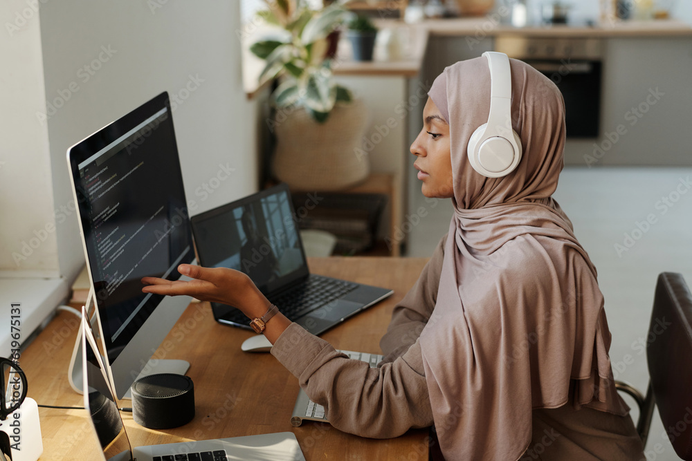 Poster Young Muslim female programmer pointing at data on computer screen while sitting by workplace and communicating with colleague in video chat