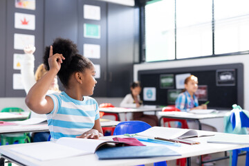 African american schoolgirl sitting at desk raising hand in elementary school classroom, copy space - obrazy, fototapety, plakaty